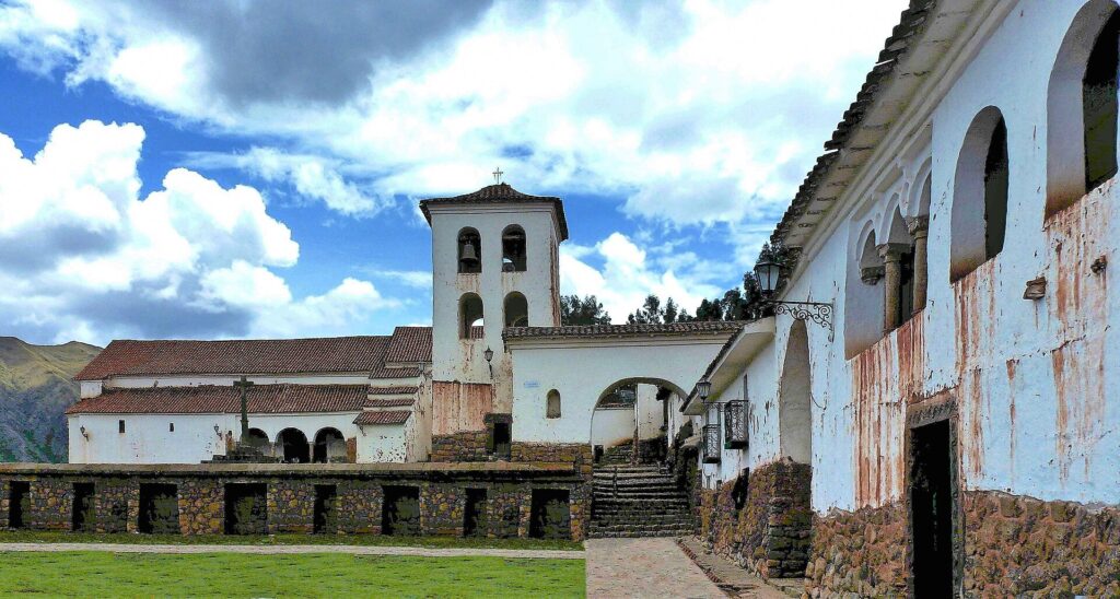 Chinchero: templo colonial blanco con campanario sobre muros incas de piedra con hornacinas, junto a una casona con arcos y un jardín verde