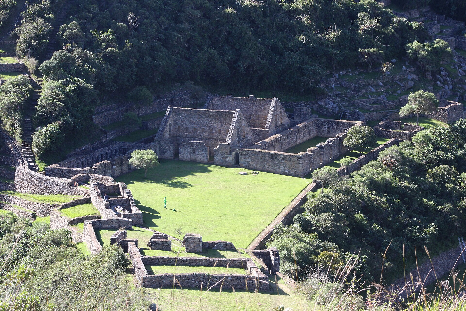 Edificios de piedra inca y una plaza de césped rodeados de bosque, vistos desde arriba en Choquequirao