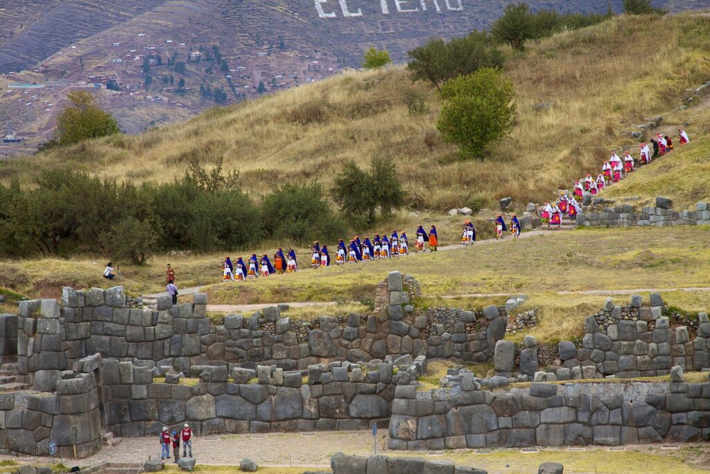 Inti Raymi: una larga fila de personas con trajes de colores camina por una ladera de pasto seco, sobre los muros de piedra de Sacsayhuamán
