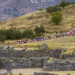 Inti Raymi: una larga fila de personas con trajes de colores camina por una ladera de pasto seco, sobre los muros de piedra de Sacsayhuamán