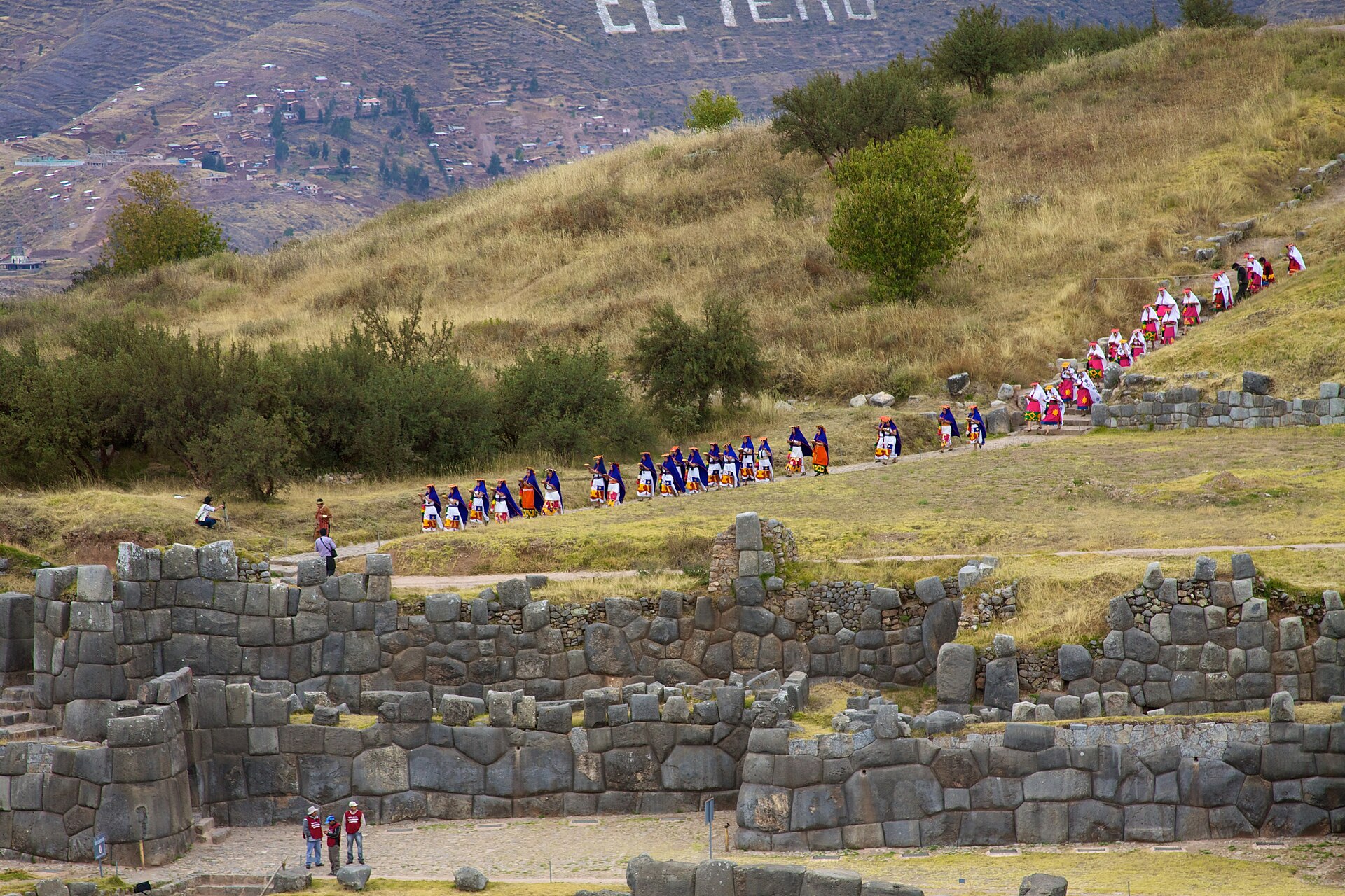Inti Raymi: una larga fila de personas con trajes de colores camina por una ladera de pasto seco, sobre los muros de piedra de Sacsayhuamán