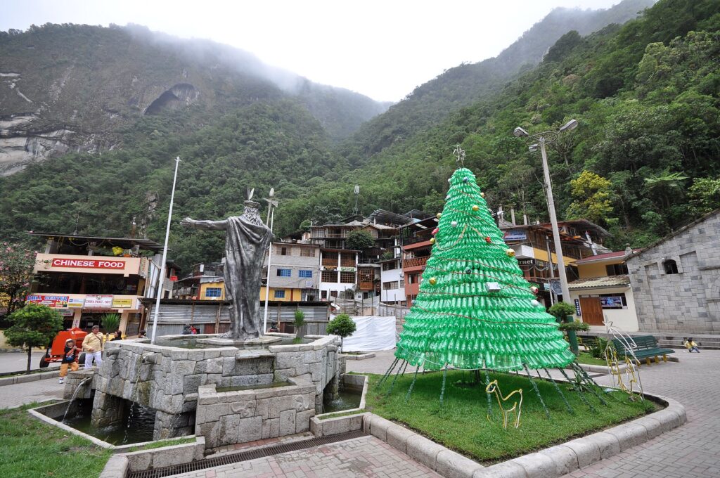 Plaza de Machupicchu Pueblo con estatua de Pachacútec, un árbol verde hecho de botellas y cerros con bosque al fondo