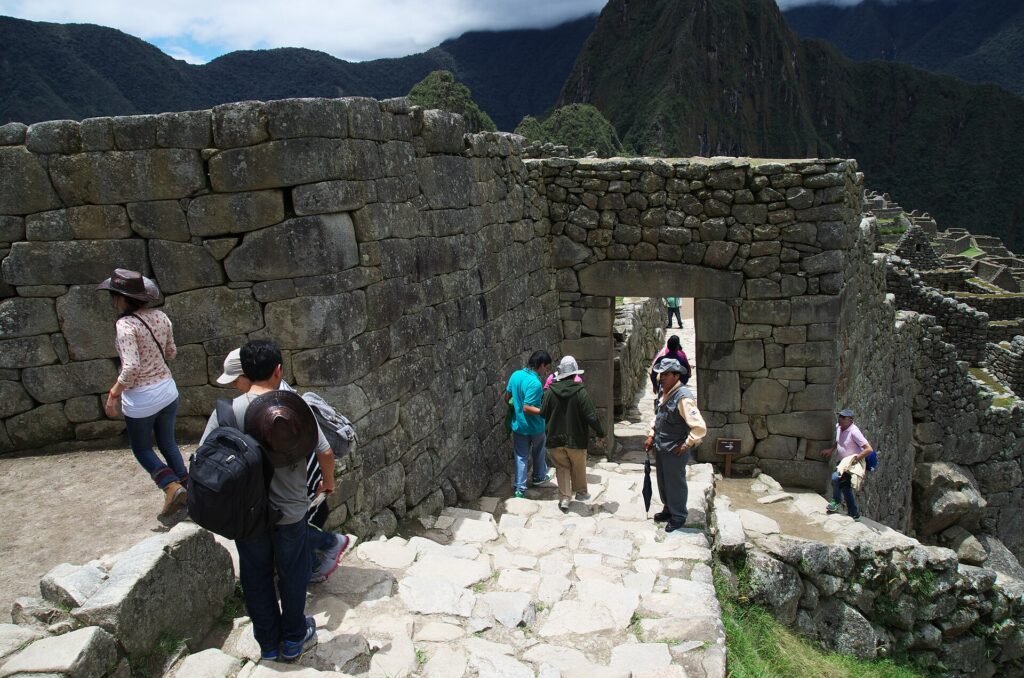 Visitantes caminan junto a muros y una portada de piedra inca en Machu Picchu, con montañas al fondo