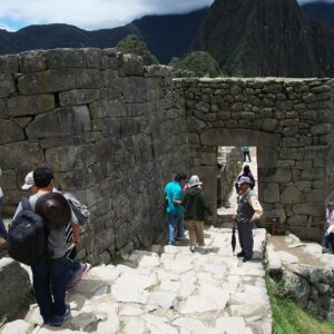 Visitantes caminan junto a muros y una portada de piedra inca en Machu Picchu, con montañas al fondo