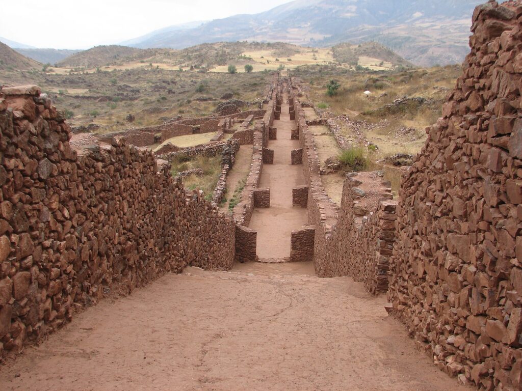 Pikillaqta: calle recta entre altos muros de piedra rojiza sin tallar que baja hacia el valle, con cerros al fondo