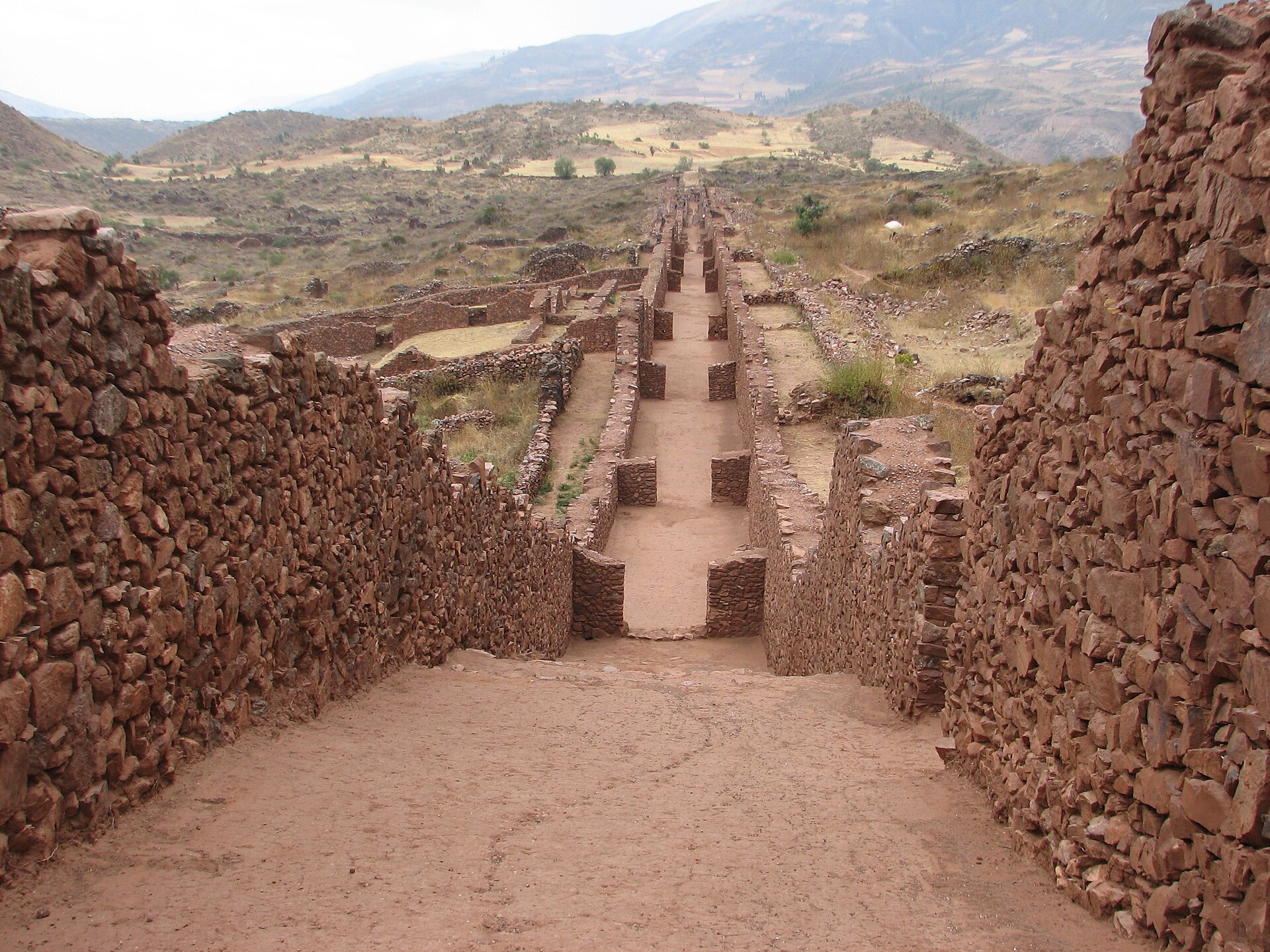 Pikillaqta: calle recta entre altos muros de piedra rojiza sin tallar que baja hacia el valle, con cerros al fondo