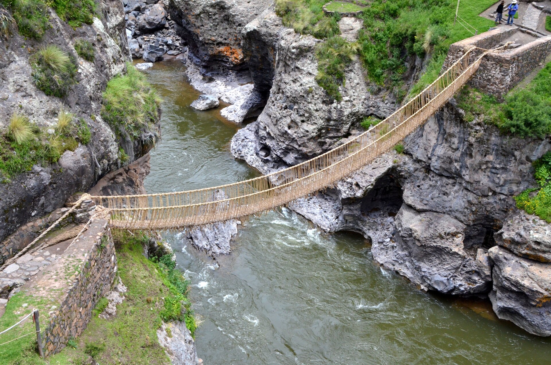 Puente Q'eswachaka: puente colgante de cuerdas de paja dorada cruza un río verde entre paredes de roca gris, con bases de piedra a cada lado