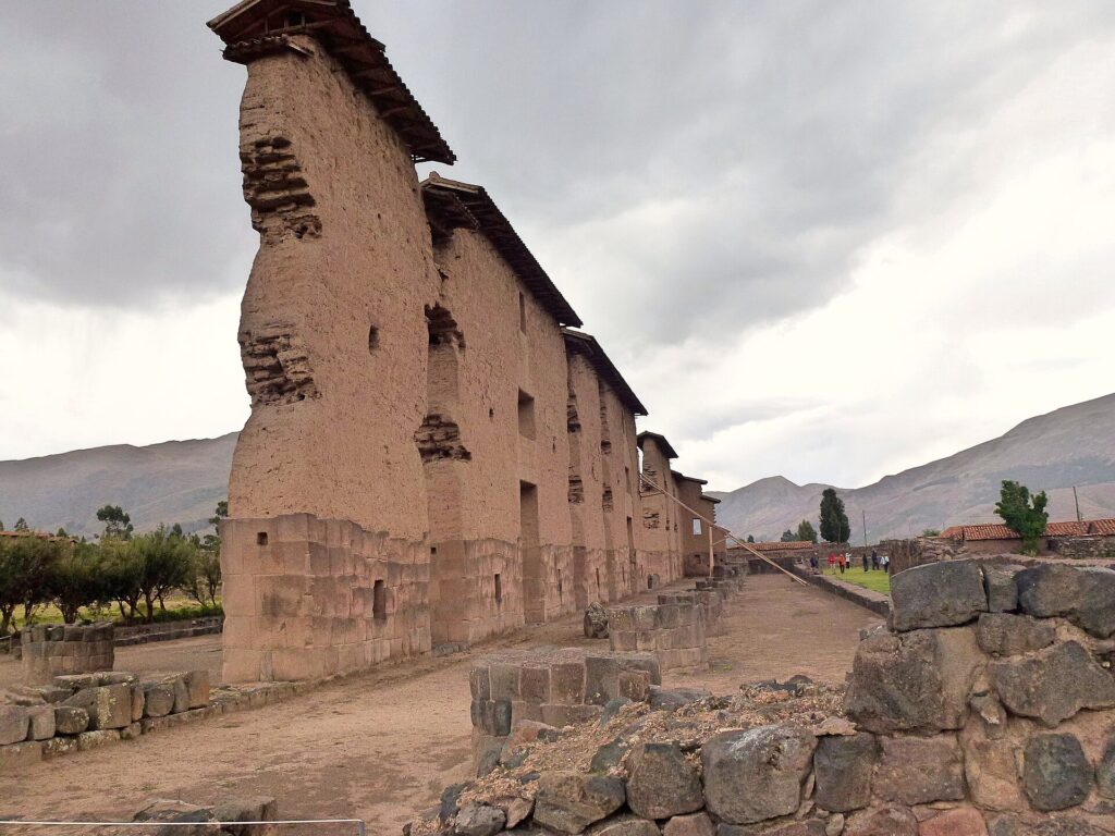 Raqchi: alto muro del templo de Viracocha con base de piedra y parte superior de adobe, con pequeños techos, junto a un camino de tierra y cerros al fondo