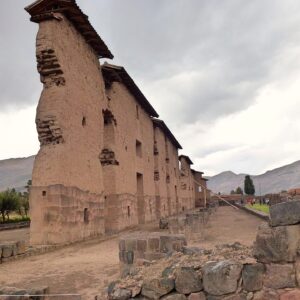 Raqchi: alto muro del templo de Viracocha con base de piedra y parte superior de adobe, con pequeños techos, junto a un camino de tierra y cerros al fondo