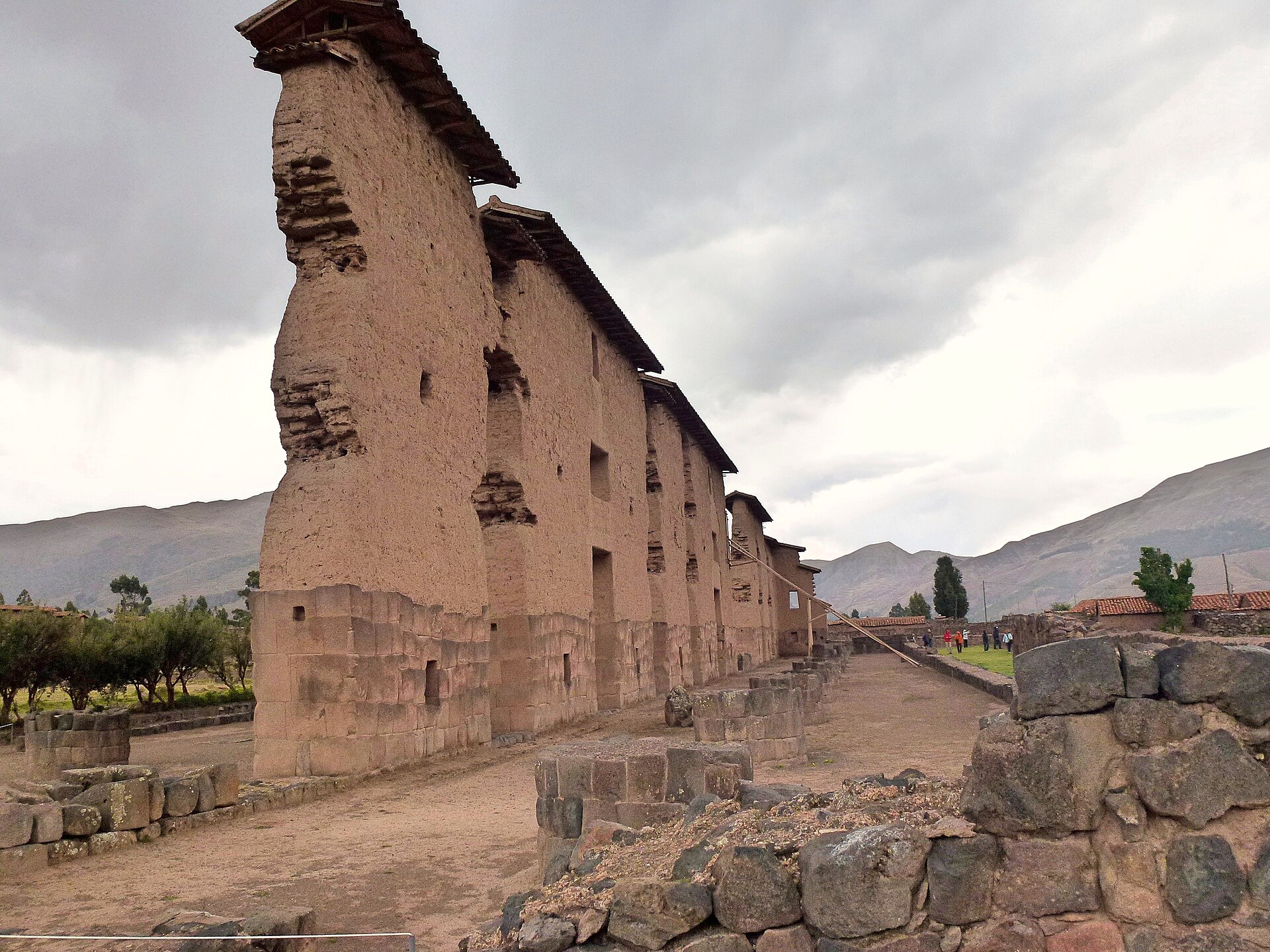Raqchi: alto muro del templo de Viracocha con base de piedra y parte superior de adobe, con pequeños techos, junto a un camino de tierra y cerros al fondo