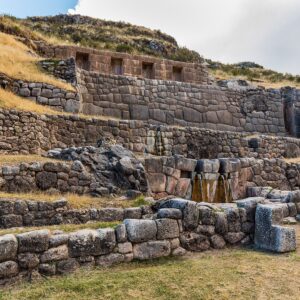 Tambomachay: terrazas incas de piedra en una ladera con pasto seco, con nichos en los muros altos y una fuente de agua al centro, bajo un cielo nublado