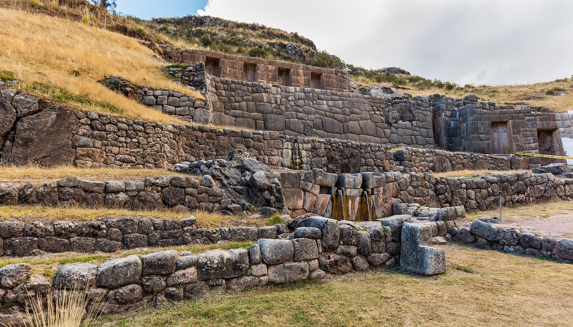 Tambomachay: terrazas incas de piedra en una ladera con pasto seco, con nichos en los muros altos y una fuente de agua al centro, bajo un cielo nublado