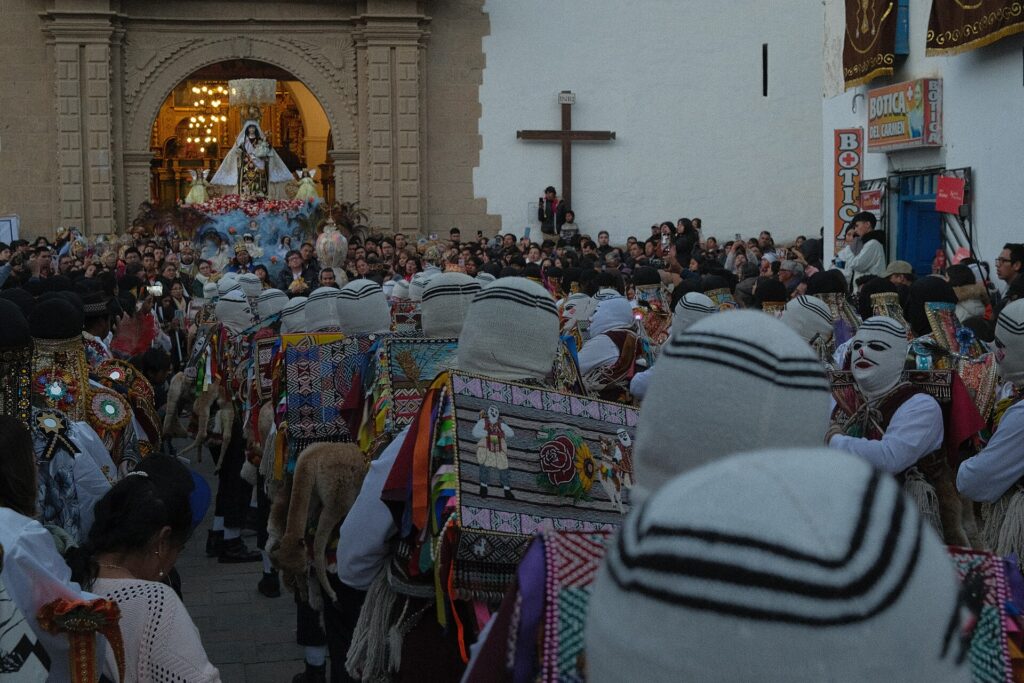 Virgen del Carmen de Paucartambo: danzantes Qhapaq Qolla con máscaras blancas y gorros tejidos frente a la portada del templo, entre una multitud