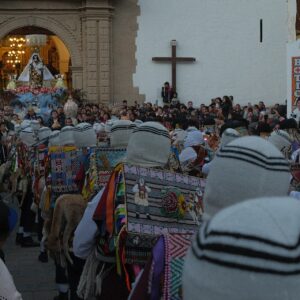 Virgen del Carmen de Paucartambo: danzantes Qhapaq Qolla con máscaras blancas y gorros tejidos frente a la portada del templo, entre una multitud