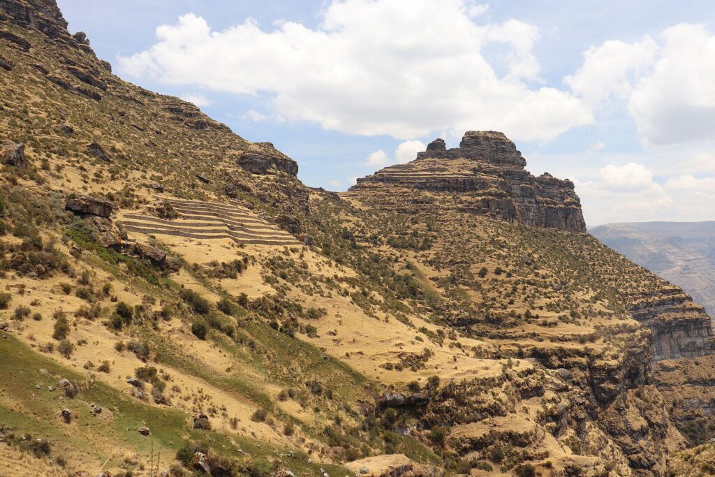 Waqrapukara: gran roca escalonada en la cima de un cerro, sobre laderas de pasto seco y arbustos, con andenes incas a la izquierda y nubes blancas