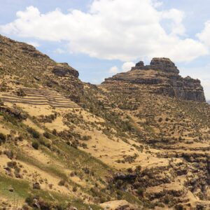 Waqrapukara: gran roca escalonada en la cima de un cerro, sobre laderas de pasto seco y arbustos, con andenes incas a la izquierda y nubes blancas
