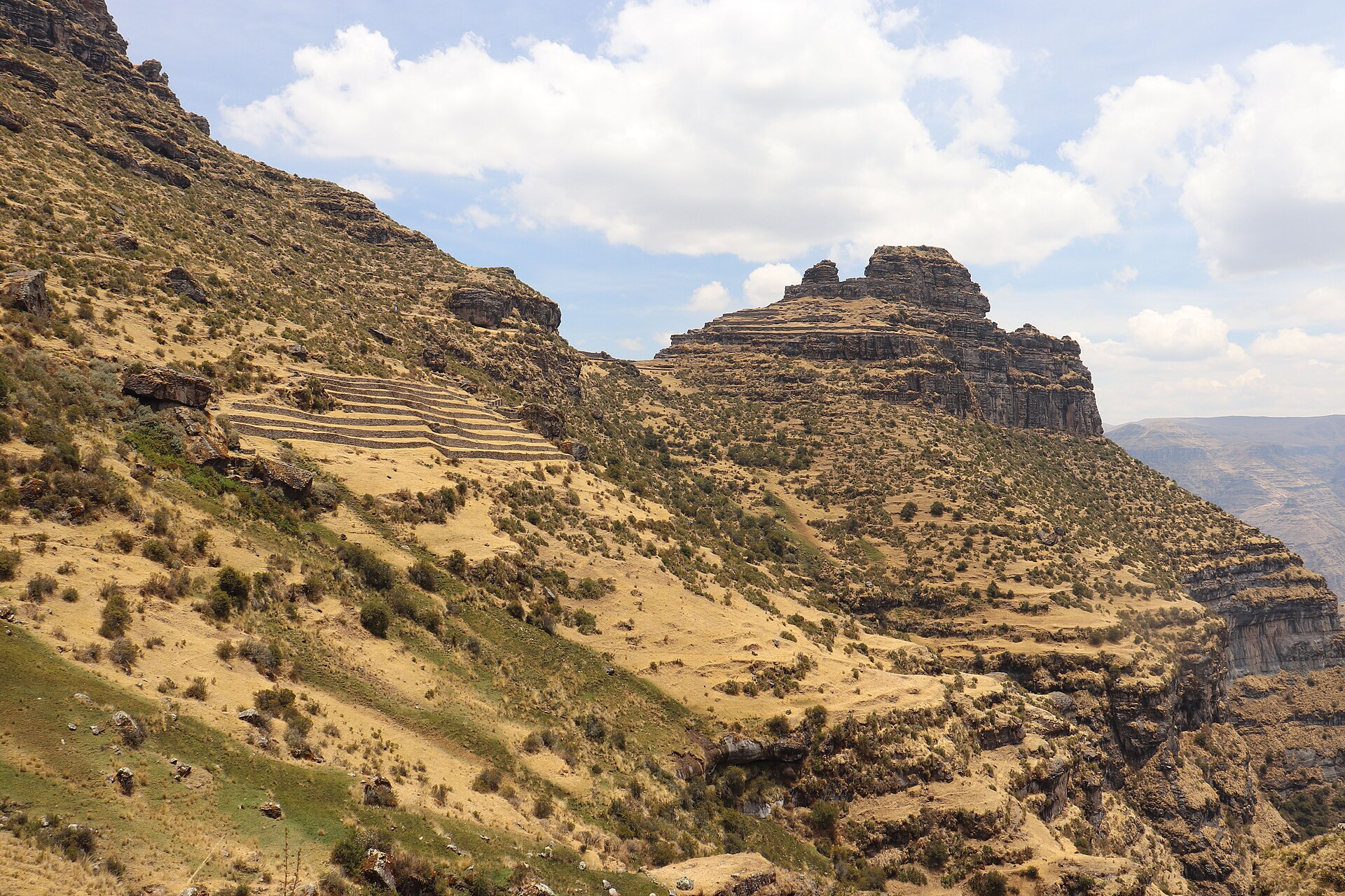 Waqrapukara: gran roca escalonada en la cima de un cerro, sobre laderas de pasto seco y arbustos, con andenes incas a la izquierda y nubes blancas